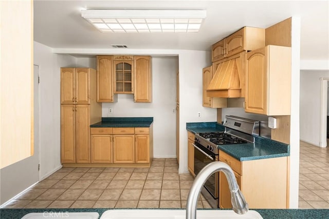 kitchen with custom exhaust hood, light tile patterned floors, dark countertops, glass insert cabinets, and stainless steel gas range oven