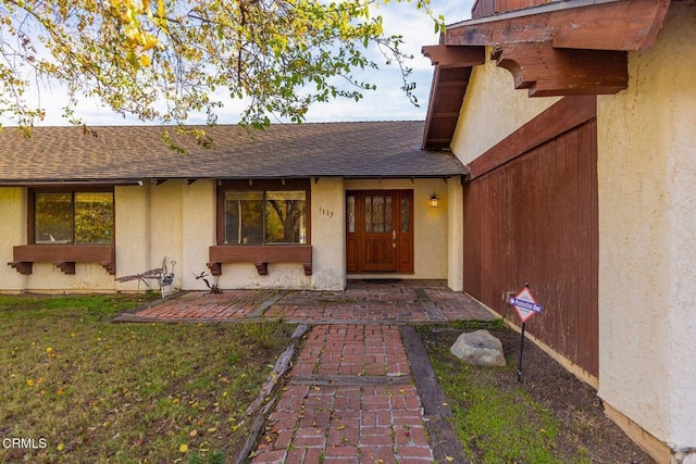 view of exterior entry featuring a yard, roof with shingles, and stucco siding