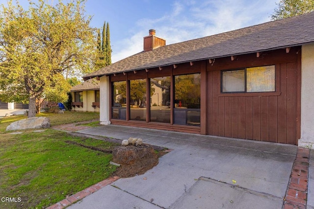 rear view of house with roof with shingles, a lawn, a chimney, and a patio