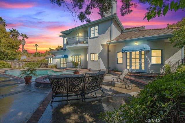 back of house at dusk featuring french doors, a patio, a balcony, and stucco siding