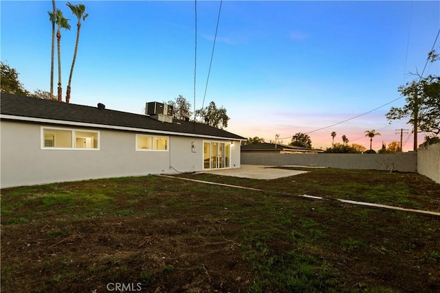 back of property at dusk with a patio, central air condition unit, fence, a yard, and stucco siding