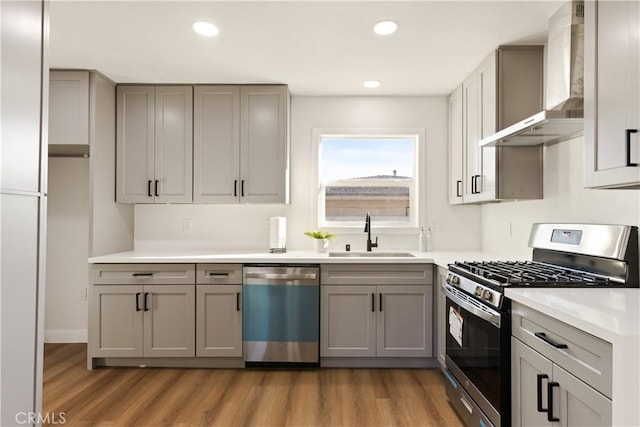 kitchen with light wood finished floors, gray cabinets, appliances with stainless steel finishes, a sink, and wall chimney range hood