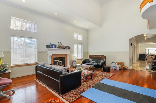 living room featuring arched walkways, a tiled fireplace, a high ceiling, ornamental molding, and wood finished floors