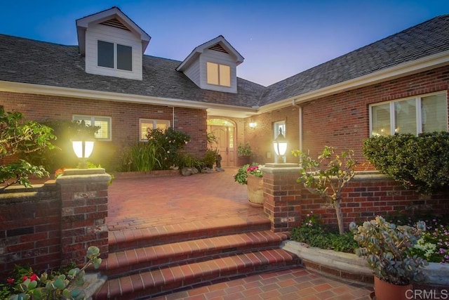 exterior entry at dusk featuring a shingled roof, brick siding, and a patio