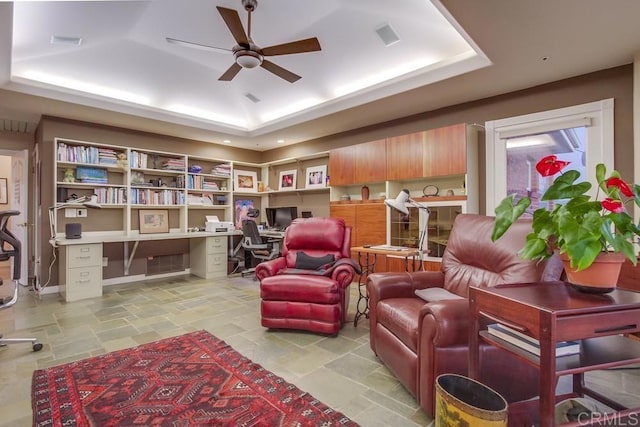 living area featuring stone finish flooring, a raised ceiling, built in study area, and a ceiling fan