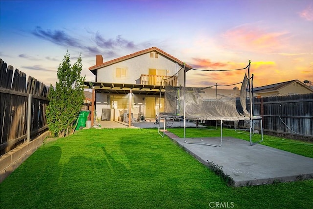 back of house at dusk with a fenced backyard, a trampoline, a lawn, and a patio