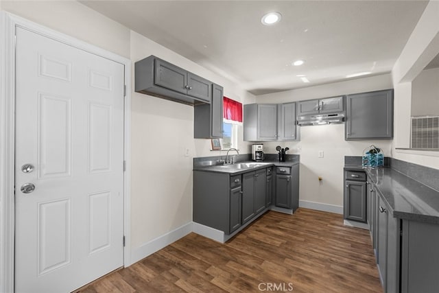 kitchen with dark wood-style floors, dark countertops, gray cabinetry, a sink, and under cabinet range hood