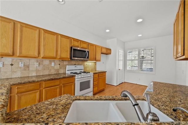 kitchen featuring white gas range oven, stainless steel microwave, dark stone countertops, a sink, and backsplash