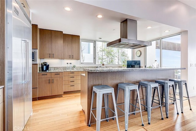 kitchen with built in refrigerator, light wood-style flooring, island exhaust hood, and a breakfast bar