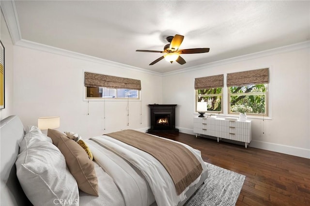 bedroom with a warm lit fireplace, dark wood-style flooring, a ceiling fan, baseboards, and crown molding