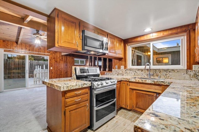 kitchen featuring brown cabinetry, a ceiling fan, a sink, stainless steel appliances, and wooden walls