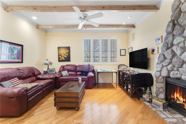 living area with beam ceiling, light wood-style floors, ornamental molding, a ceiling fan, and a stone fireplace