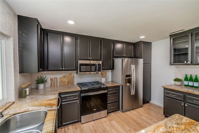 kitchen with stainless steel appliances, a sink, light wood-style floors, light stone countertops, and glass insert cabinets