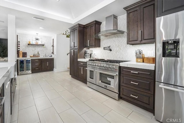 kitchen featuring appliances with stainless steel finishes, light countertops, dark brown cabinetry, and wall chimney exhaust hood