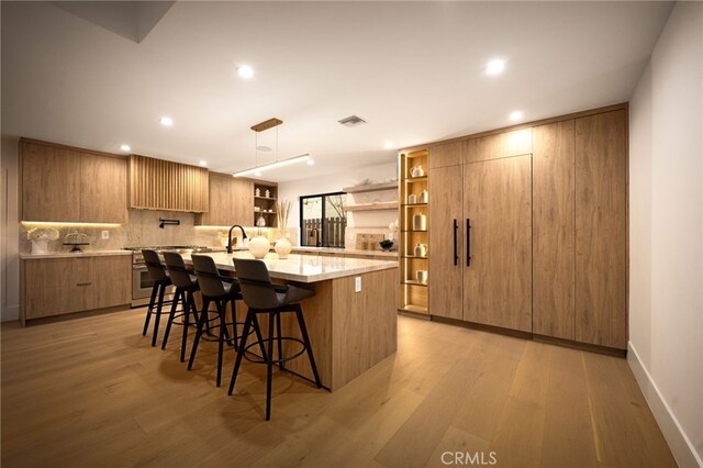 kitchen featuring a kitchen island with sink, decorative backsplash, open shelves, light wood finished floors, and a kitchen bar