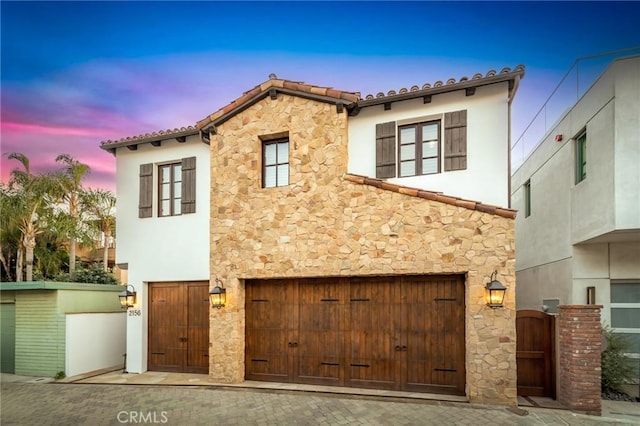 mediterranean / spanish-style home featuring a garage, a tile roof, stone siding, a gate, and stucco siding