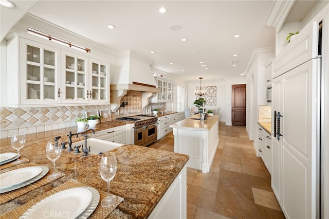 kitchen with a sink, custom exhaust hood, double oven range, a center island with sink, and crown molding