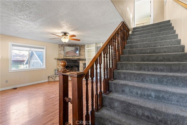 stairs with a textured ceiling, a stone fireplace, wood finished floors, a ceiling fan, and baseboards