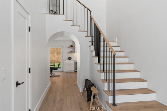 stairs featuring wine cooler, arched walkways, a towering ceiling, wood finished floors, and baseboards