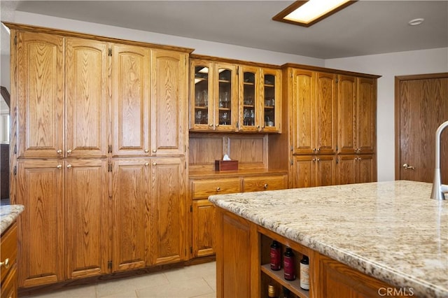 kitchen featuring light stone counters, glass insert cabinets, brown cabinets, and light tile patterned floors