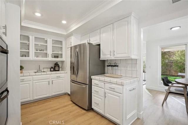 kitchen with stainless steel fridge, white cabinets, tile countertops, glass insert cabinets, and crown molding