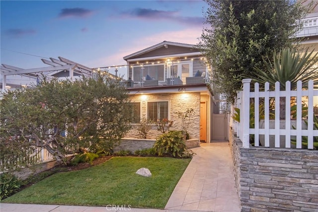 view of front of house with a balcony, stone siding, fence, and a front yard