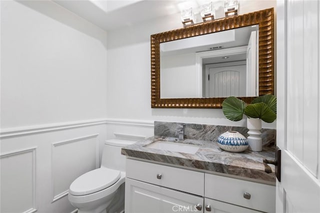 bathroom featuring a wainscoted wall, vanity, toilet, and a decorative wall