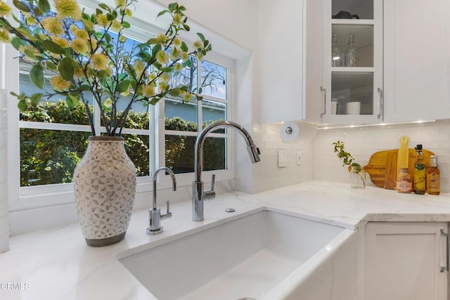 interior details featuring white cabinets, glass insert cabinets, a sink, light stone countertops, and backsplash