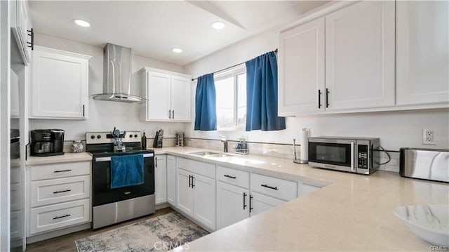 kitchen with light countertops, appliances with stainless steel finishes, white cabinetry, a sink, and wall chimney range hood