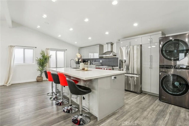 kitchen featuring stacked washer and clothes dryer, an island with sink, a breakfast bar area, wall chimney range hood, and stainless steel refrigerator with ice dispenser