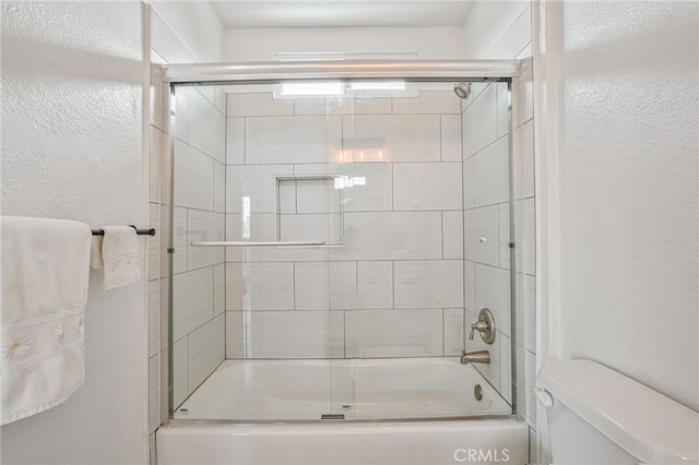 full bath featuring toilet, enclosed tub / shower combo, and a textured wall