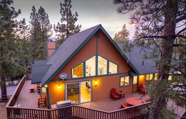 back of house at dusk featuring a deck, a chimney, and a shingled roof