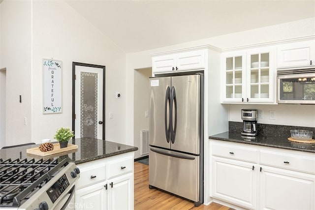 kitchen featuring stainless steel appliances, vaulted ceiling, white cabinets, glass insert cabinets, and dark stone countertops