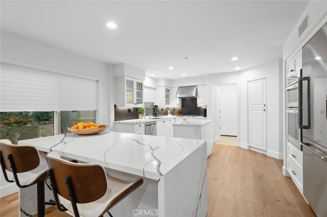 kitchen with stainless steel appliances, wall chimney range hood, light wood-type flooring, and glass insert cabinets