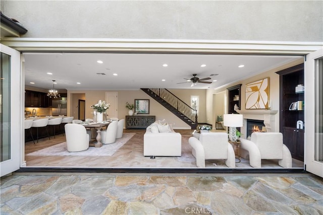 living room featuring ceiling fan with notable chandelier, recessed lighting, ornamental molding, a glass covered fireplace, and stairs