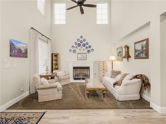 living area featuring wood finished floors, a glass covered fireplace, a ceiling fan, and baseboards