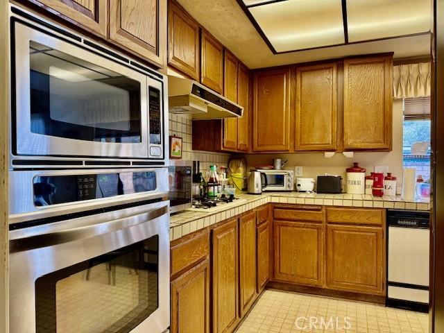 kitchen with appliances with stainless steel finishes, brown cabinetry, tile counters, and under cabinet range hood