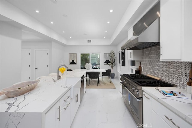 kitchen with light stone counters, white cabinetry, wall chimney range hood, decorative backsplash, and a tray ceiling