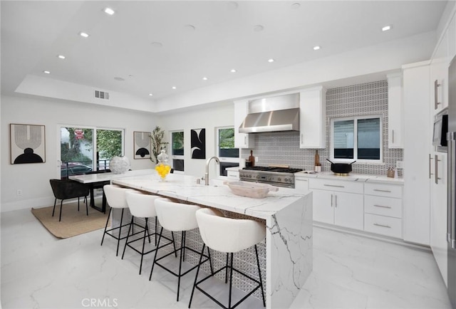 kitchen featuring a kitchen island with sink, visible vents, high end stainless steel range, wall chimney exhaust hood, and a tray ceiling