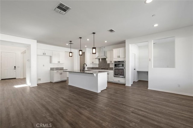 kitchen featuring dark wood-style flooring, visible vents, stainless steel double oven, a kitchen island with sink, and wall chimney exhaust hood