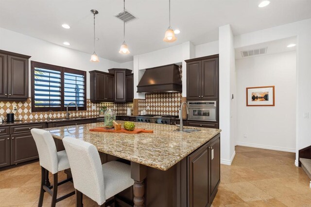 kitchen with a center island with sink, custom exhaust hood, visible vents, stainless steel oven, and dark brown cabinets