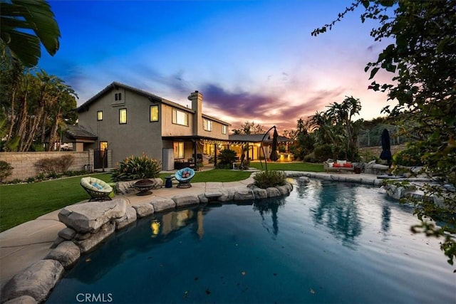pool at dusk featuring a lawn, fence, an outdoor pool, an outdoor fire pit, and a patio area