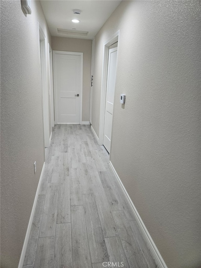 corridor featuring a textured wall, light wood-type flooring, and baseboards