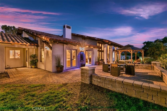 back of house at dusk with a patio, a chimney, a tiled roof, a gazebo, and stucco siding