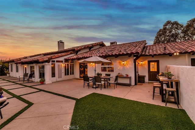 back of property at dusk featuring a patio, a chimney, a tiled roof, a yard, and stucco siding