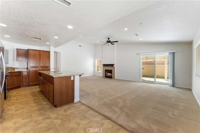 kitchen featuring a textured ceiling, light stone counters, light colored carpet, brown cabinets, and a tiled fireplace