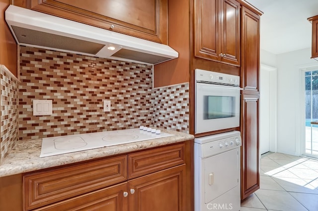 kitchen featuring white appliances, tasteful backsplash, brown cabinetry, light stone counters, and under cabinet range hood