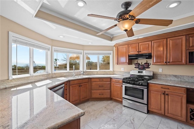 kitchen with a tray ceiling, a sink, under cabinet range hood, appliances with stainless steel finishes, and brown cabinets