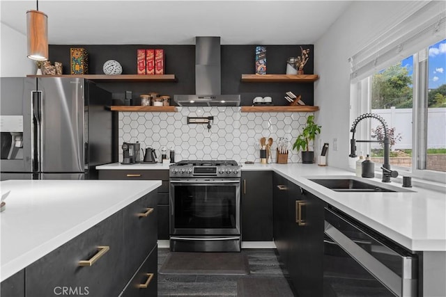 kitchen with appliances with stainless steel finishes, dark cabinetry, wall chimney range hood, open shelves, and a sink