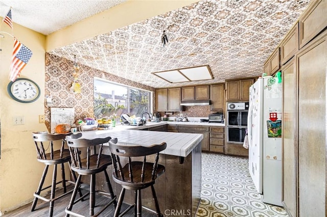 kitchen with freestanding refrigerator, stainless steel double oven, a peninsula, under cabinet range hood, and a kitchen breakfast bar